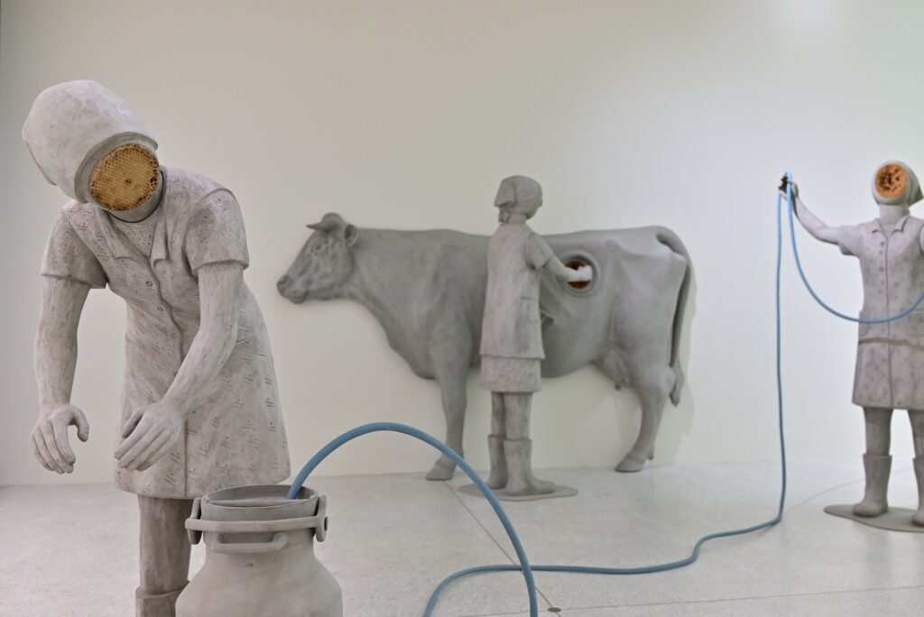Drei graue Arbeiterinnenfiguren mit Wabeneinsätzen, Kuh, Milchkanne und blauem Schlauch in Anna Hulačovás After Bugonia.