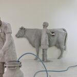 Drei graue Arbeiterinnenfiguren mit Wabeneinsätzen, Kuh, Milchkanne und blauem Schlauch in Anna Hulačovás After Bugonia.