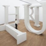 Monumentale weiße Betonbuchstaben von Shilpa Guptas Truth stehen versetzt; ein T liegt am Boden, ein Mensch zwischen den Lettern verdeutlicht ihre Größe.