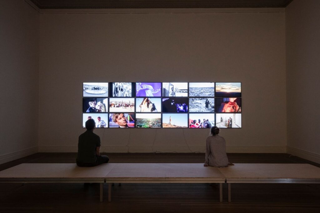 Zwei Personen sitzen vor einer Wand mit Videobildschirmen der Installation The African Twin Towers von Christoph Schlingensief im Gropius Bau.