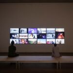 Zwei Personen sitzen vor einer Wand mit Videobildschirmen der Installation The African Twin Towers von Christoph Schlingensief im Gropius Bau.